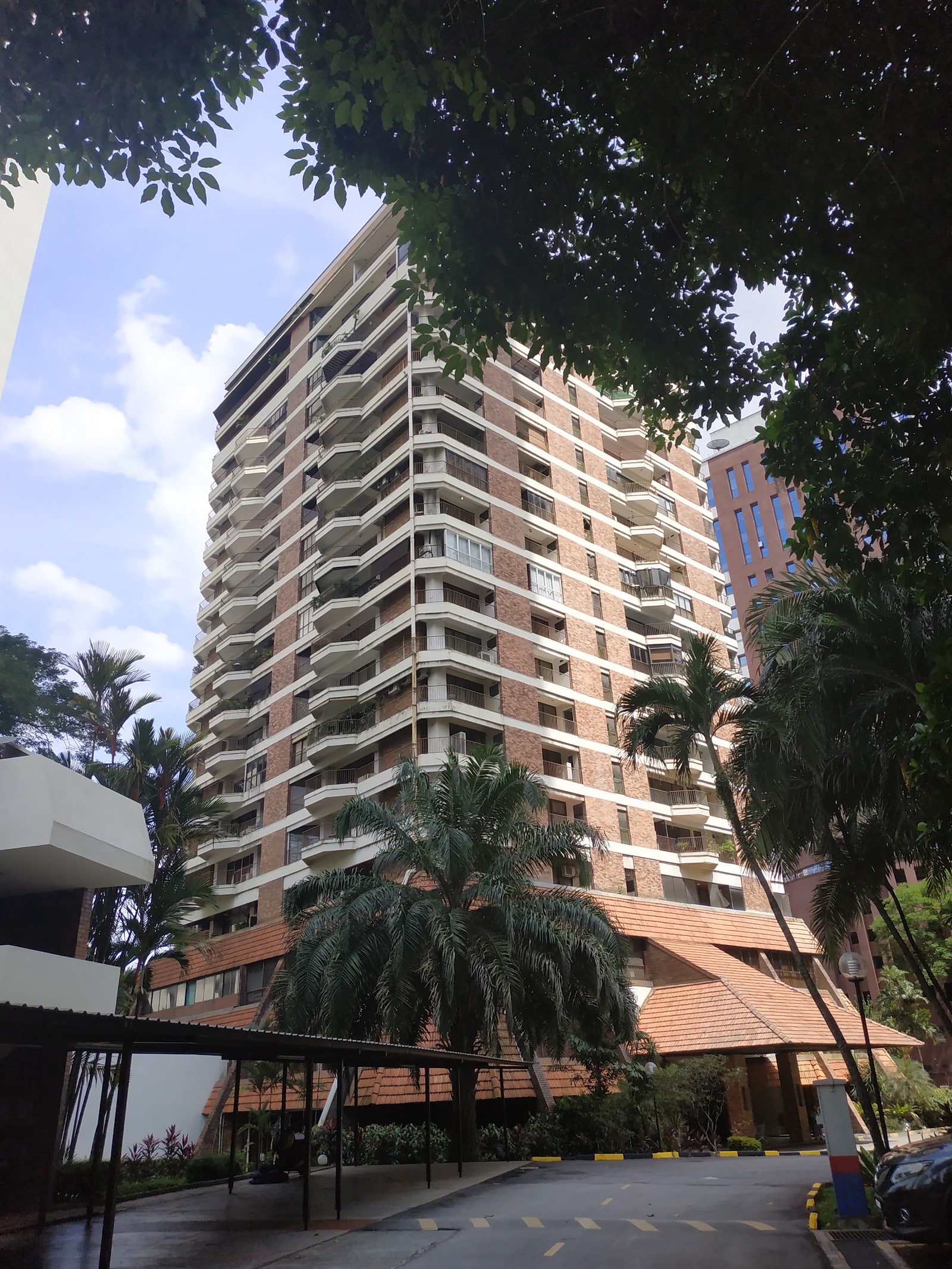 Desa Kudalari: A tall, terracotta brick and white concrete residential tower with curved balconies, viewed from the ground level surrounded by tropical trees and a paved driveway.