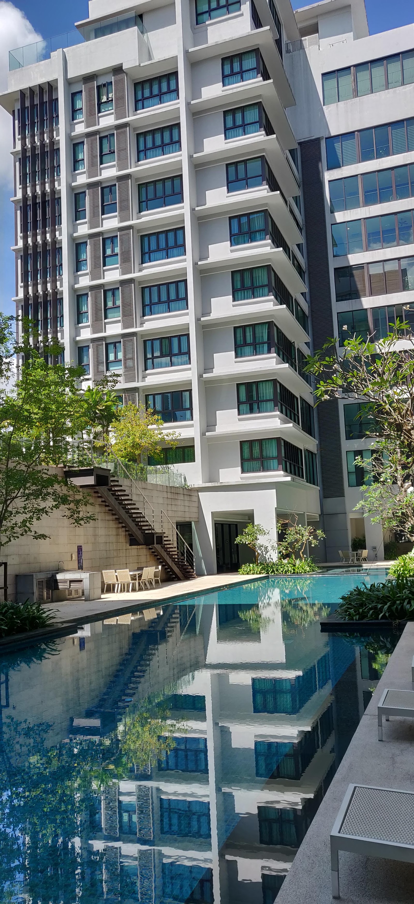 Sastra U-Thant: A swimming pool in the foreground reflecting the white facade of a residential building and the blue sky, with outdoor seating and stairs on the side.