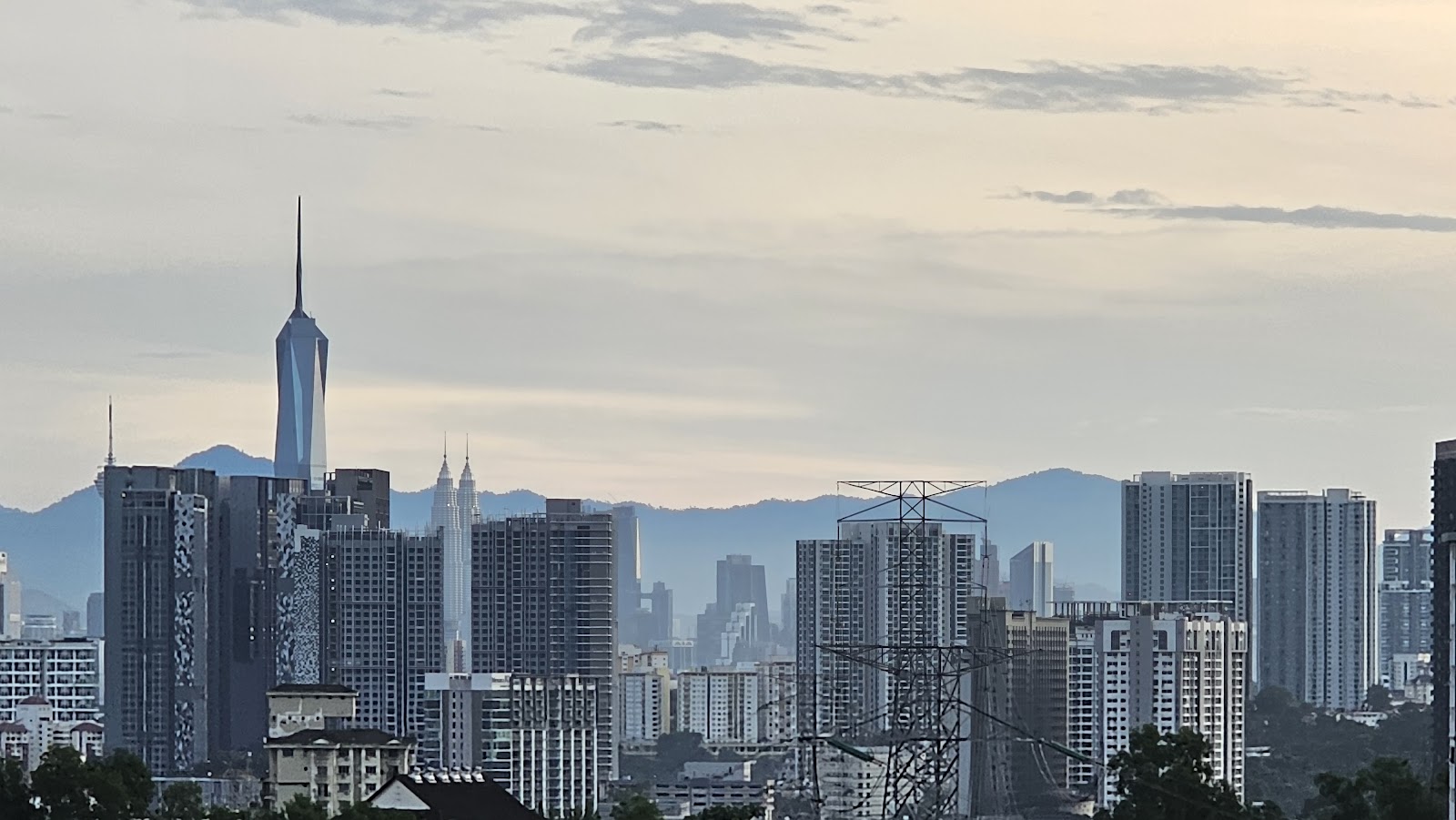 Vista OUG: A wide cityscape view from a high vantage point, showing a skyline of modern buildings, including the Petronas Twin Towers, set against hazy mountains.