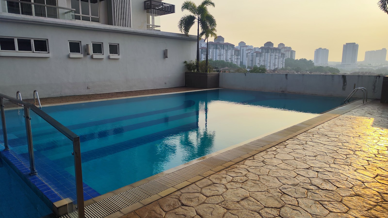 Residensi Bukit Jalil: A rooftop swimming pool with a metal ladder and stone paving, overlooking a hazy city skyline at dusk.