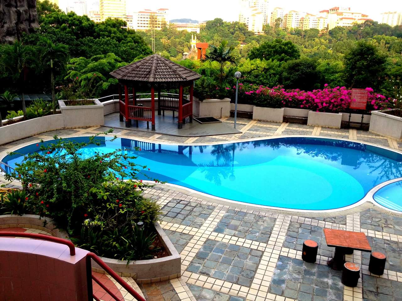 Indah Villa: An outdoor swimming pool with a gazebo and lush tropical landscaping, set against a backdrop of city buildings.