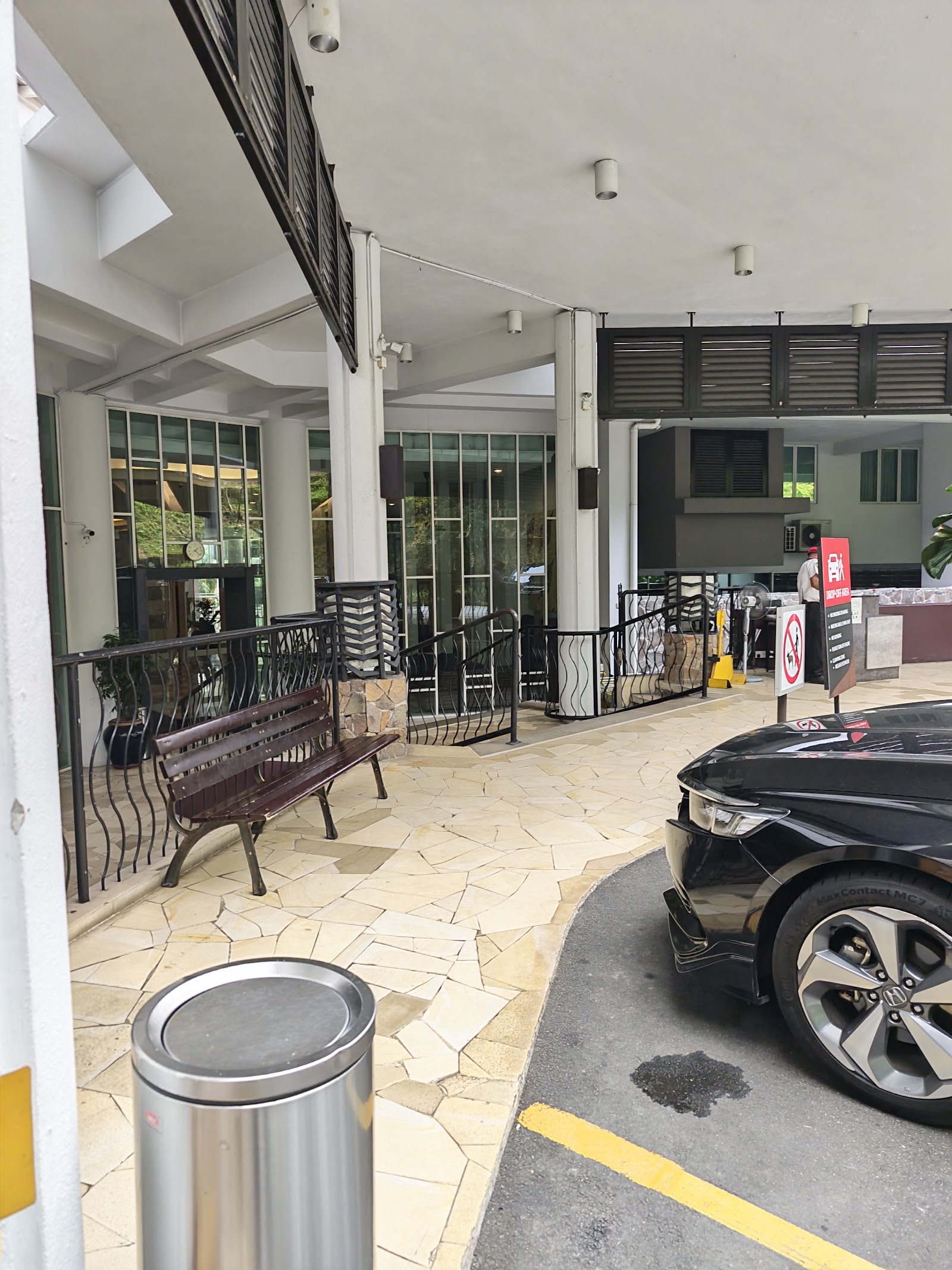 Armanee Terrace: A covered walkway area featuring a wooden bench, tiled flooring, black metal railings, a car drop-off zone with a black sedan, and a red informational sign.