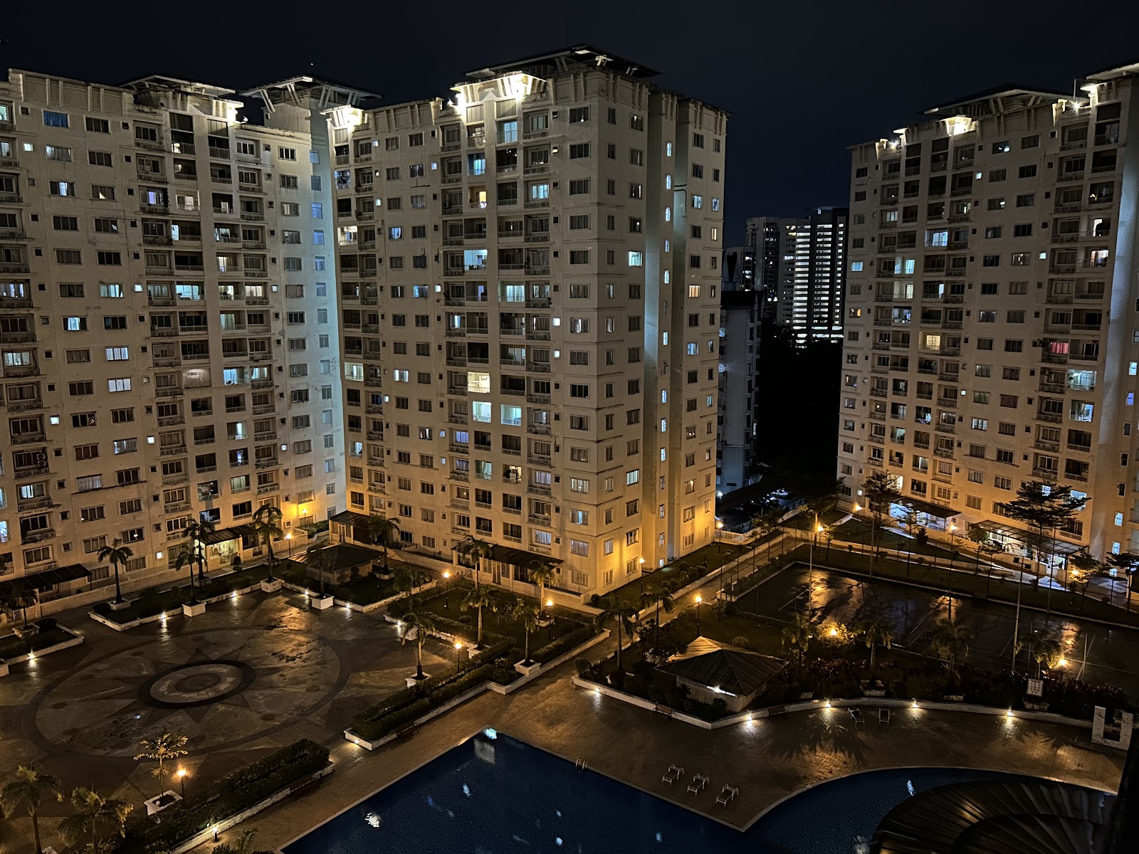 Suriamas: A night view of a residential apartment complex featuring several high-rise towers, an outdoor swimming pool, landscaped gardens, and a central courtyard with a paved area and illuminated ground.