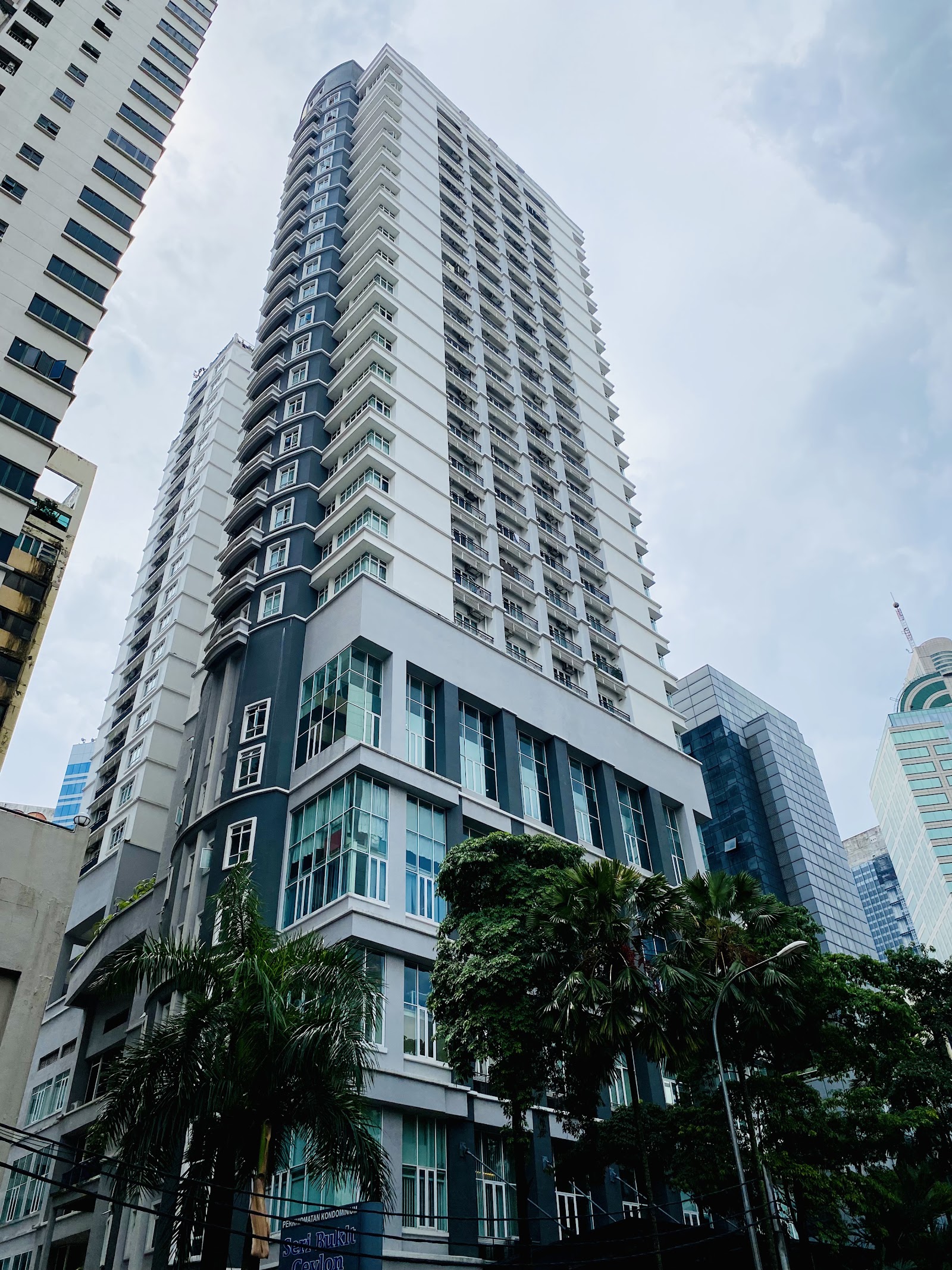Seri Bukit Ceylon: A view looking up at the exterior facade of a modern high-rise apartment building, featuring white and dark grey panels and numerous balconies, with trees at its base.