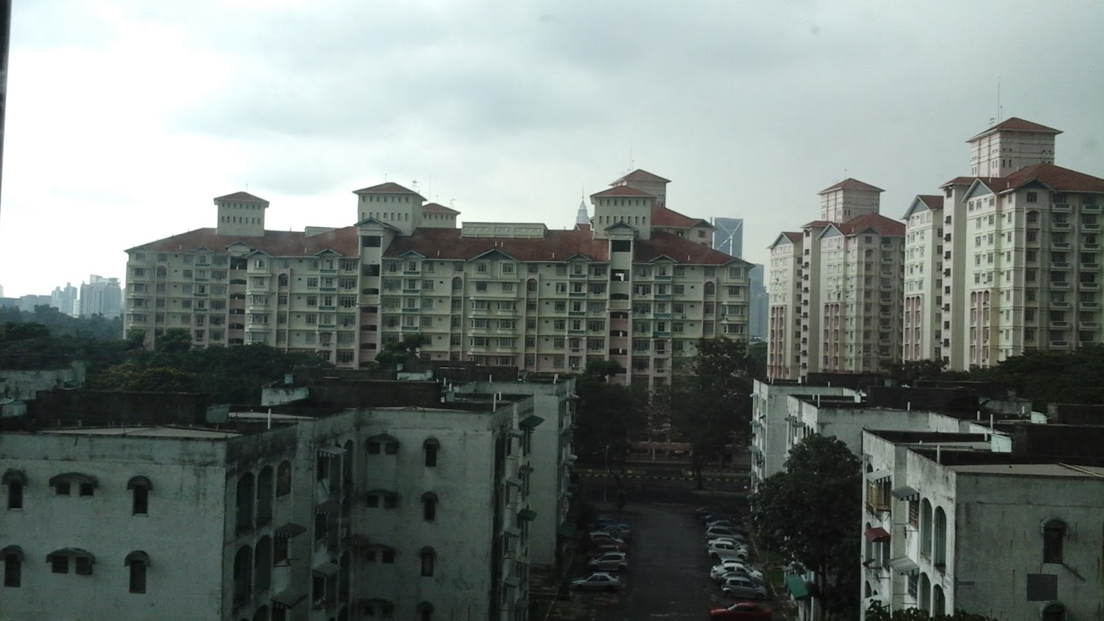 Mutiara Upper East: A wide shot from a high vantage point looking down a street lined with older residential blocks in the foreground towards large modern apartment complexes with red-tiled roofs in the distance under a cloudy sky.