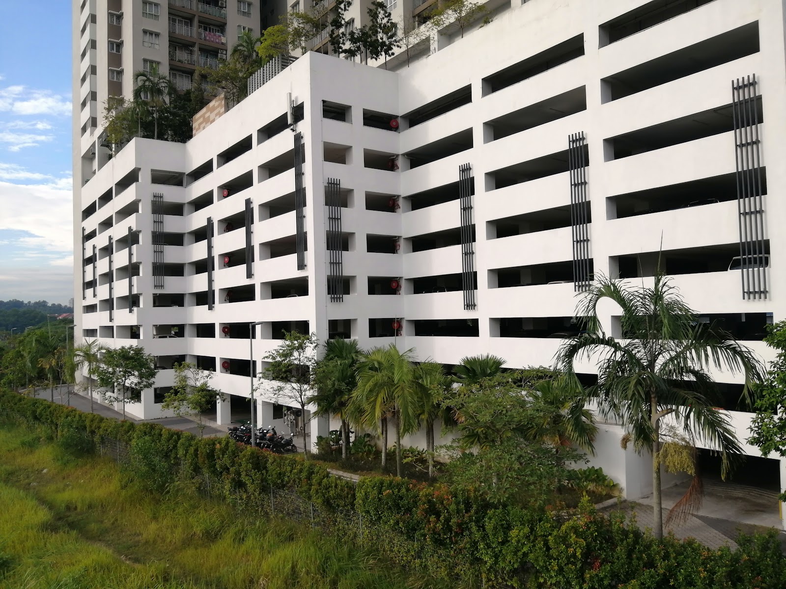 Residensi Puchongmas: The multi-story exterior of a white residential parking structure with dark vertical fins and surrounding greenery, with a high-rise tower visible in the background.