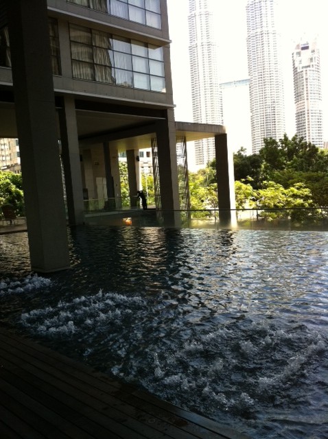 Park Seven: An outdoor swimming pool with a bubbler jet is situated under the open ground floor of a residential building, with modern high-rise towers visible in the background.