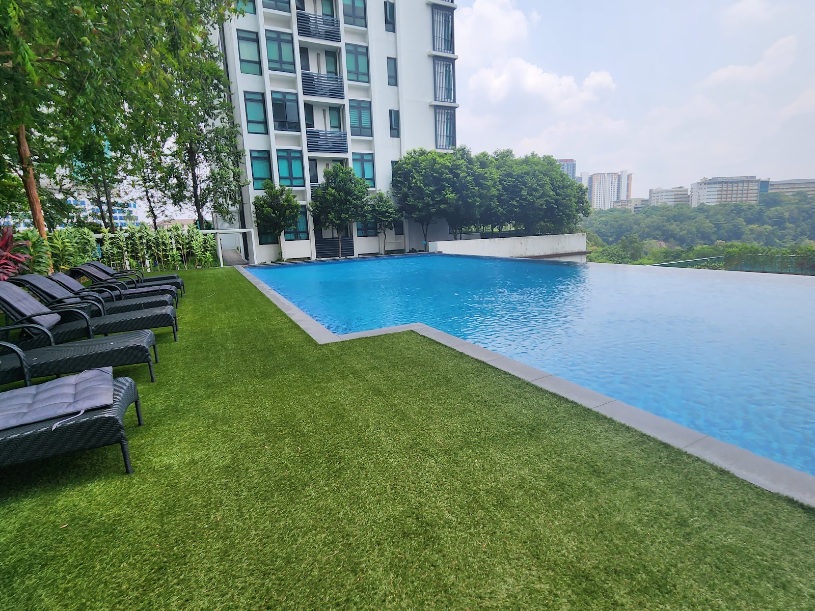 LaCosta, SUNWAY SOUTH QUAY: An outdoor rooftop infinity pool with a row of sun loungers on artificial grass, situated next to a residential building tower.