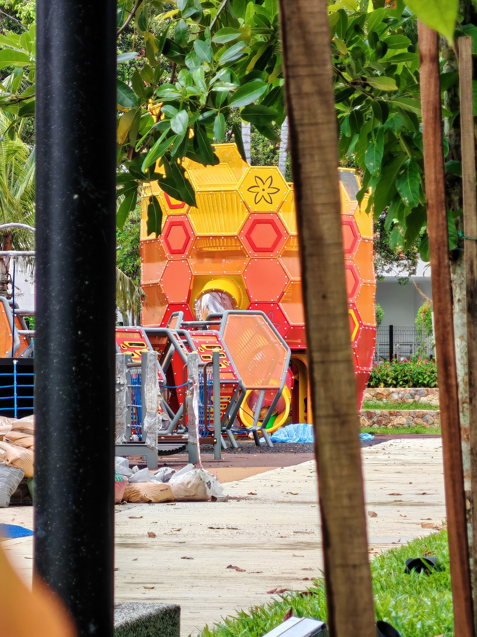 SUNWAY FLORA RESIDENCES: A view through a foreground pole and tree trunks, revealing a colorful hexagonal playground playset and some construction materials on the ground.