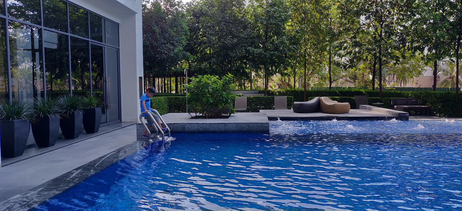 Rimbun @ Embassy Row: A swimming pool area with a person climbing up the ladder, surrounded by lounge chairs, potted plants, and a glass-walled building.