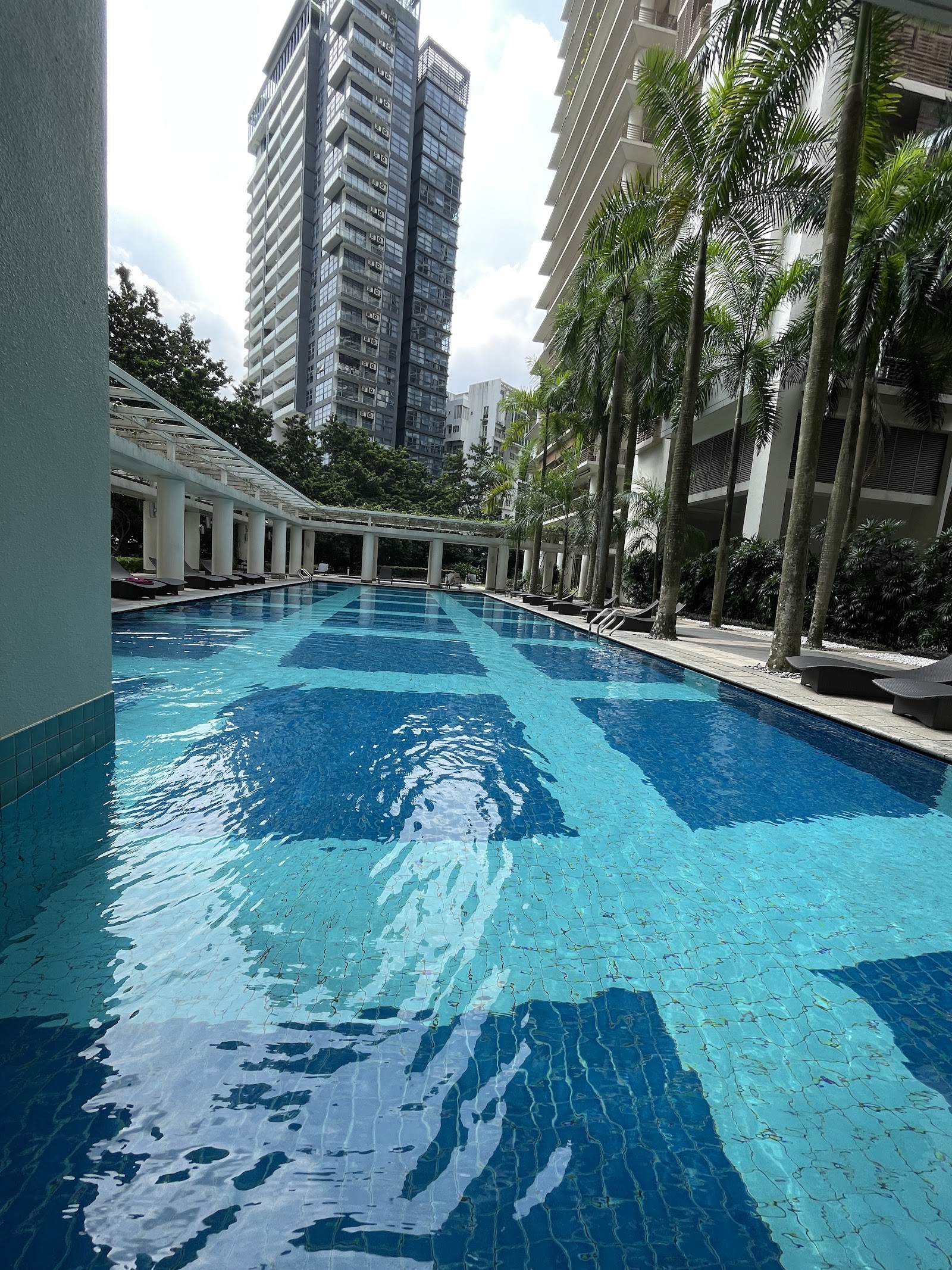 Hampshire Park: A long rectangular swimming pool in a landscaped courtyard, flanked by palm trees and a colonnade with sun loungers, with a residential tower in the background.