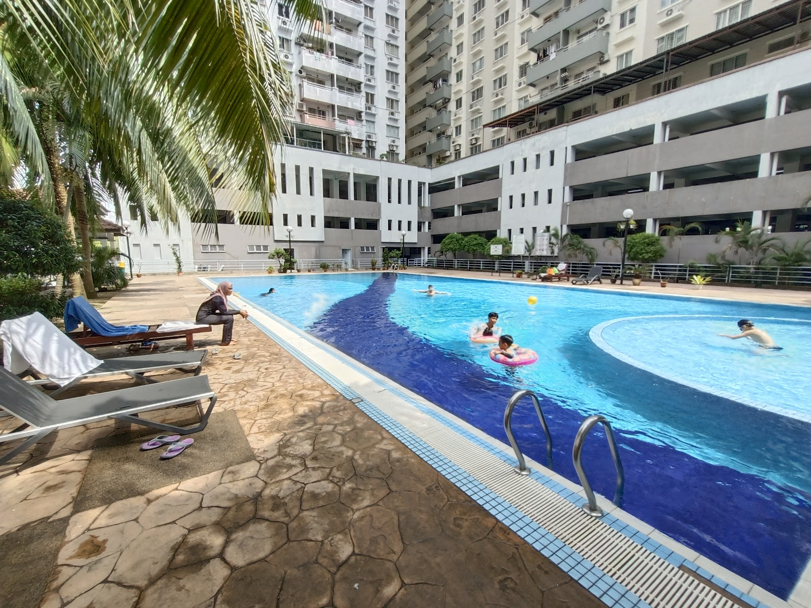 Kinrara Mas: A swimming pool within a condominium complex with residents swimming and loungers on the paved deck, surrounded by apartment towers and parking structures.