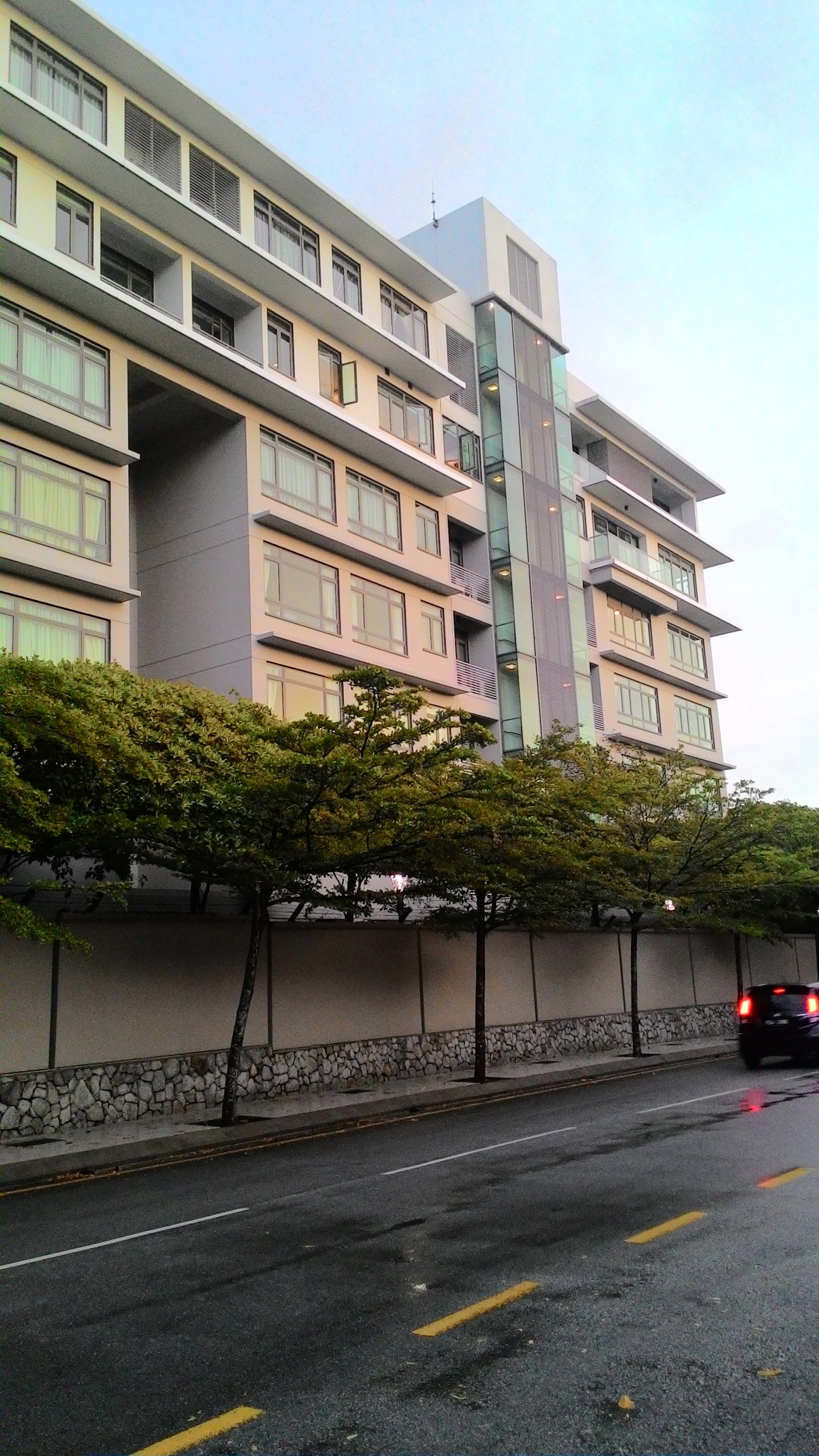 Sastra U-Thant: A street-level view of a modern residential building with a facade featuring windows, balconies, and vertical lighting accents, seen across a road and a row of trees.