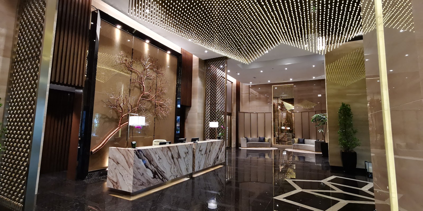 The Ritz-Carlton Residences, Kuala Lumpur: The hotel-style lobby of the residence, featuring a large marble reception desk, a striking metallic tree sculpture on the wall behind it, and a seating area in the background, all under a decorative ceiling.