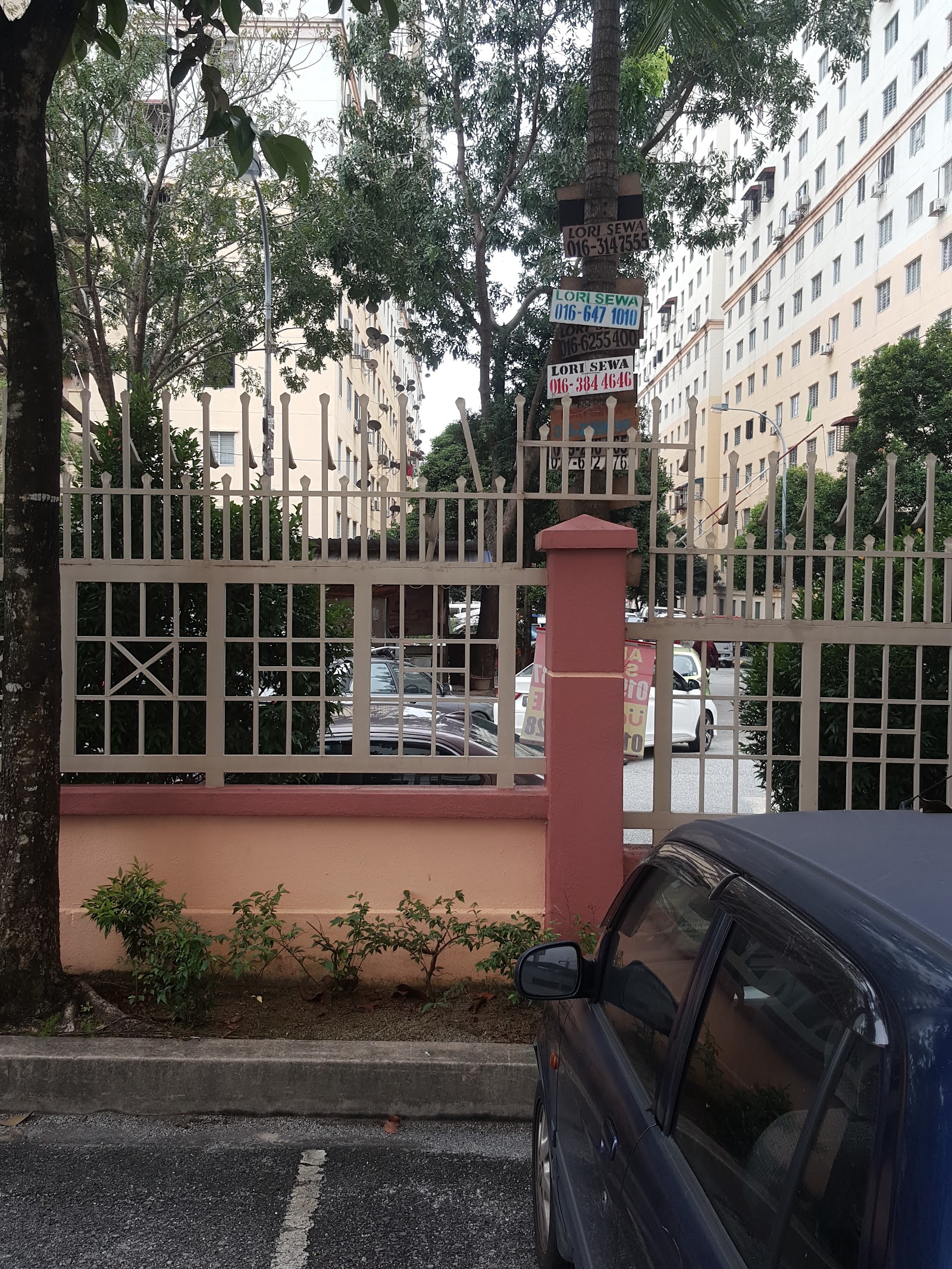 Pangsapuri Sri Rakyat: A view from the street showing a perimeter fence with a gate pillar, behind which are parked cars and high-rise residential towers in the background.