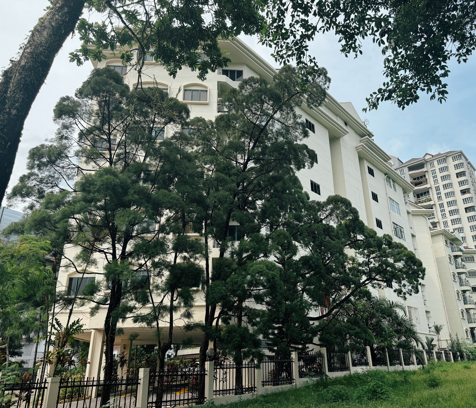 Wickham Manor: A large white residential building is partially obscured by tall trees, with a black metal fence and a patch of grass in the foreground.