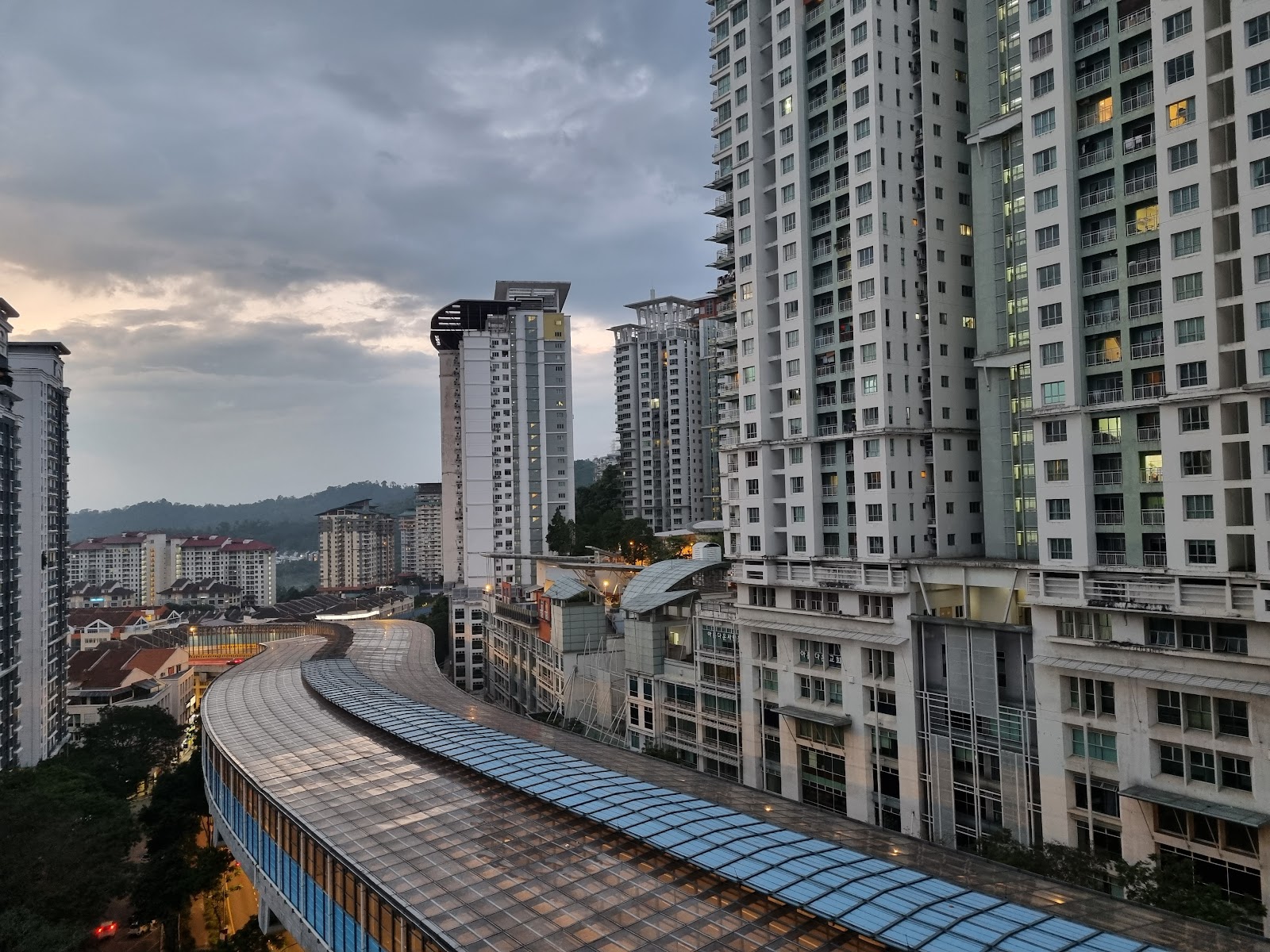 Ritze Perdana 2: A wide-angle view from a high vantage point looking out over a cityscape with high-rise buildings, a curved transit station or walkway, and distant hills under a cloudy sky.