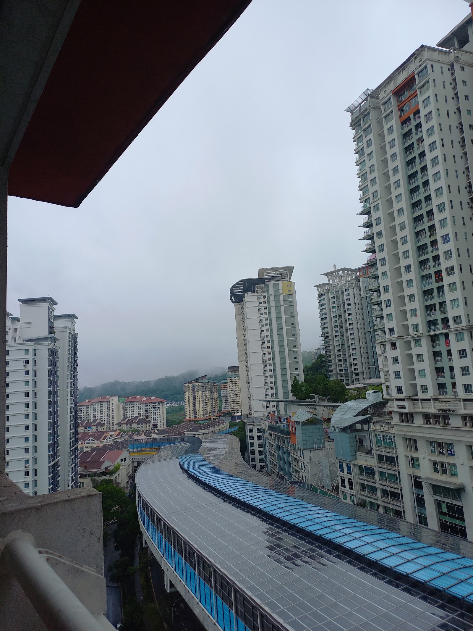 Ritze Perdana 2: A view from a high-floor balcony showing a covered elevated walkway with a blue roof, surrounding residential towers, and distant hills in a hazy atmosphere.