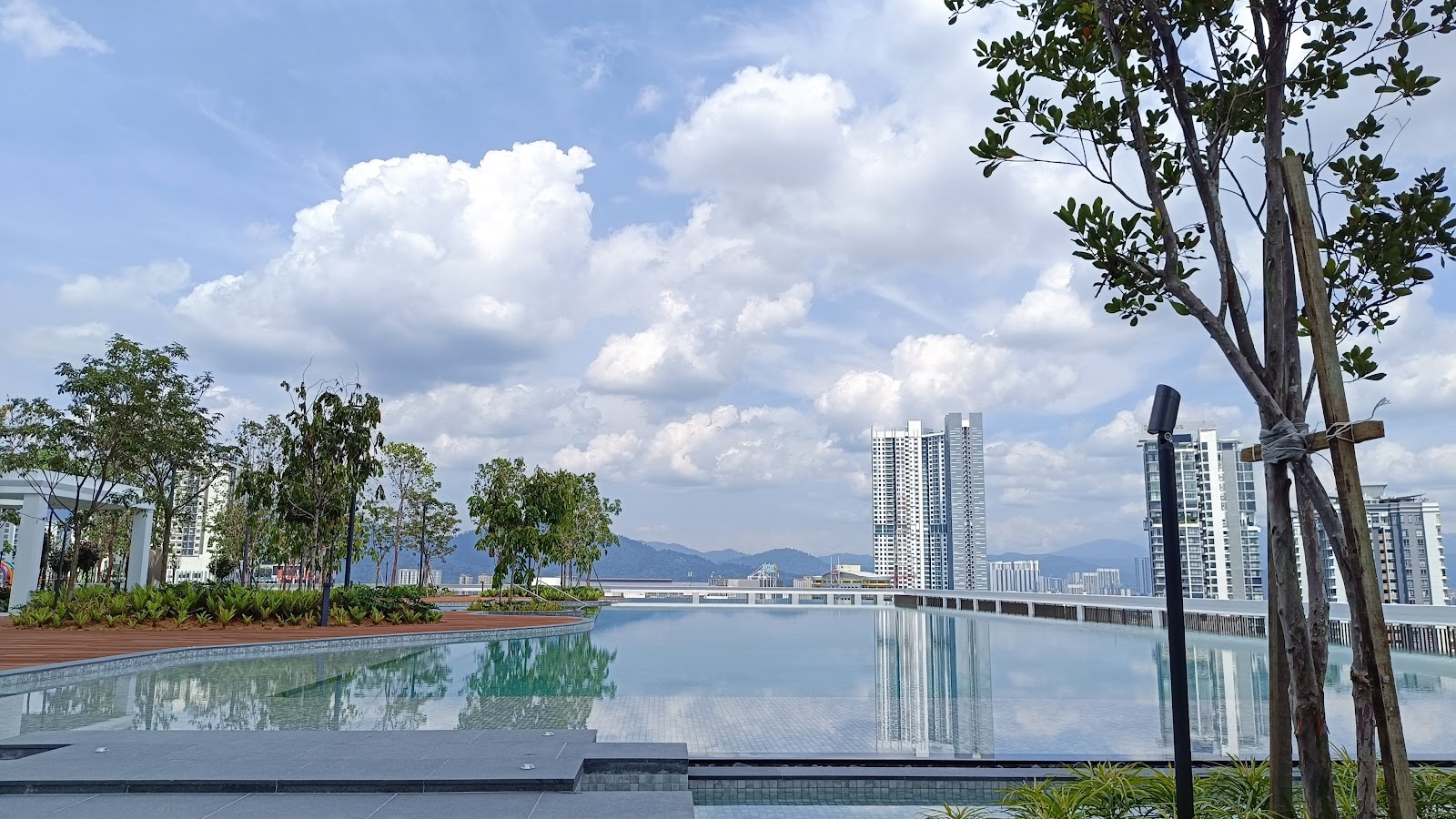 Park Place: An outdoor swimming pool deck with young trees and landscaping, overlooking a city skyline with high-rise apartments and distant mountains under a cloudy sky.