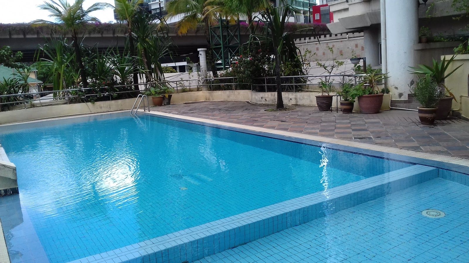Jelita Court: An outdoor swimming pool with blue tiles surrounded by a paved deck, potted plants, and a perimeter fence.