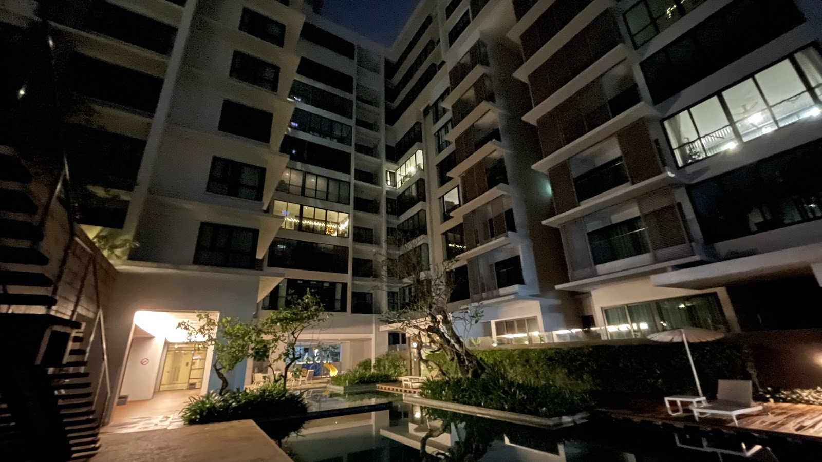 Sastra U-Thant: An outdoor swimming pool area with lounge chairs and tropical landscaping at the base of a residential building at night.