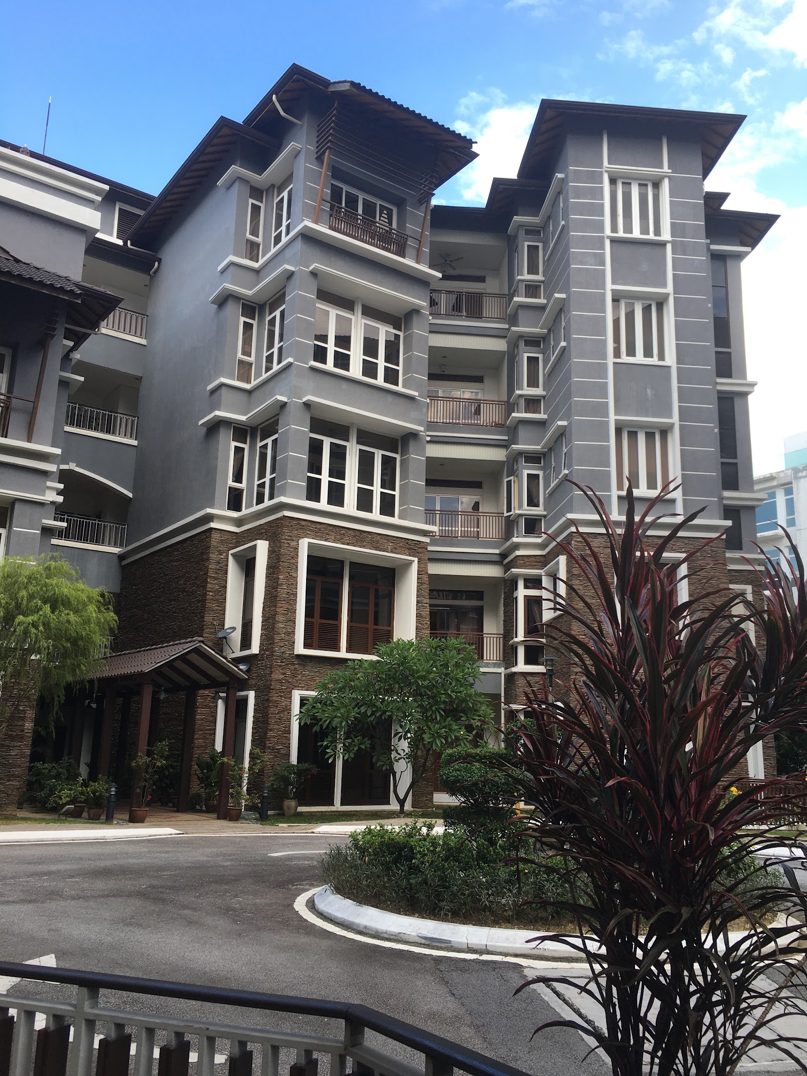 Cinta: The exterior facade of a multi-story residential building featuring grey and stone-textured finishes, white window frames, and balconies, surrounded by landscaping including green trees and a red-leafed plant in the foreground.