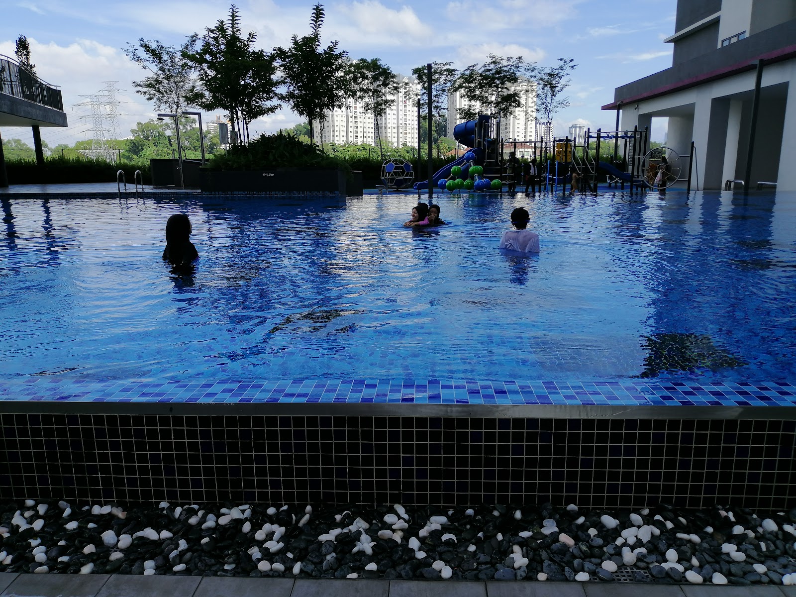 Casa Green: A swimming pool area with children swimming, featuring a blue water slide and play equipment in the background and a modern building on the right.