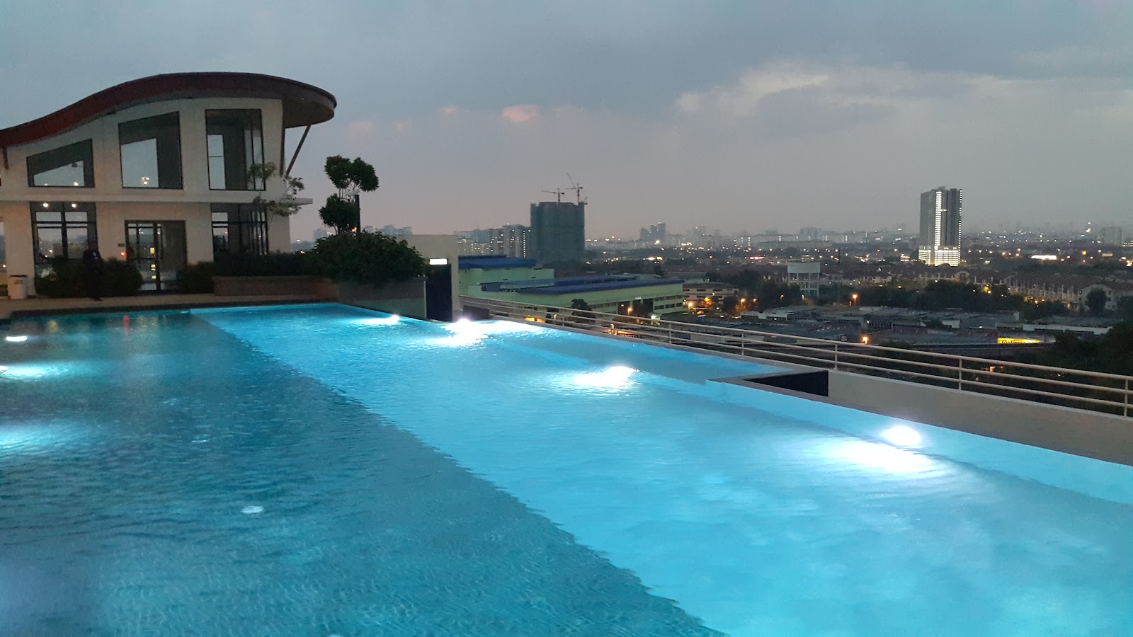 Denai Sutera: An illuminated rooftop swimming pool at dusk with a city skyline in the background.