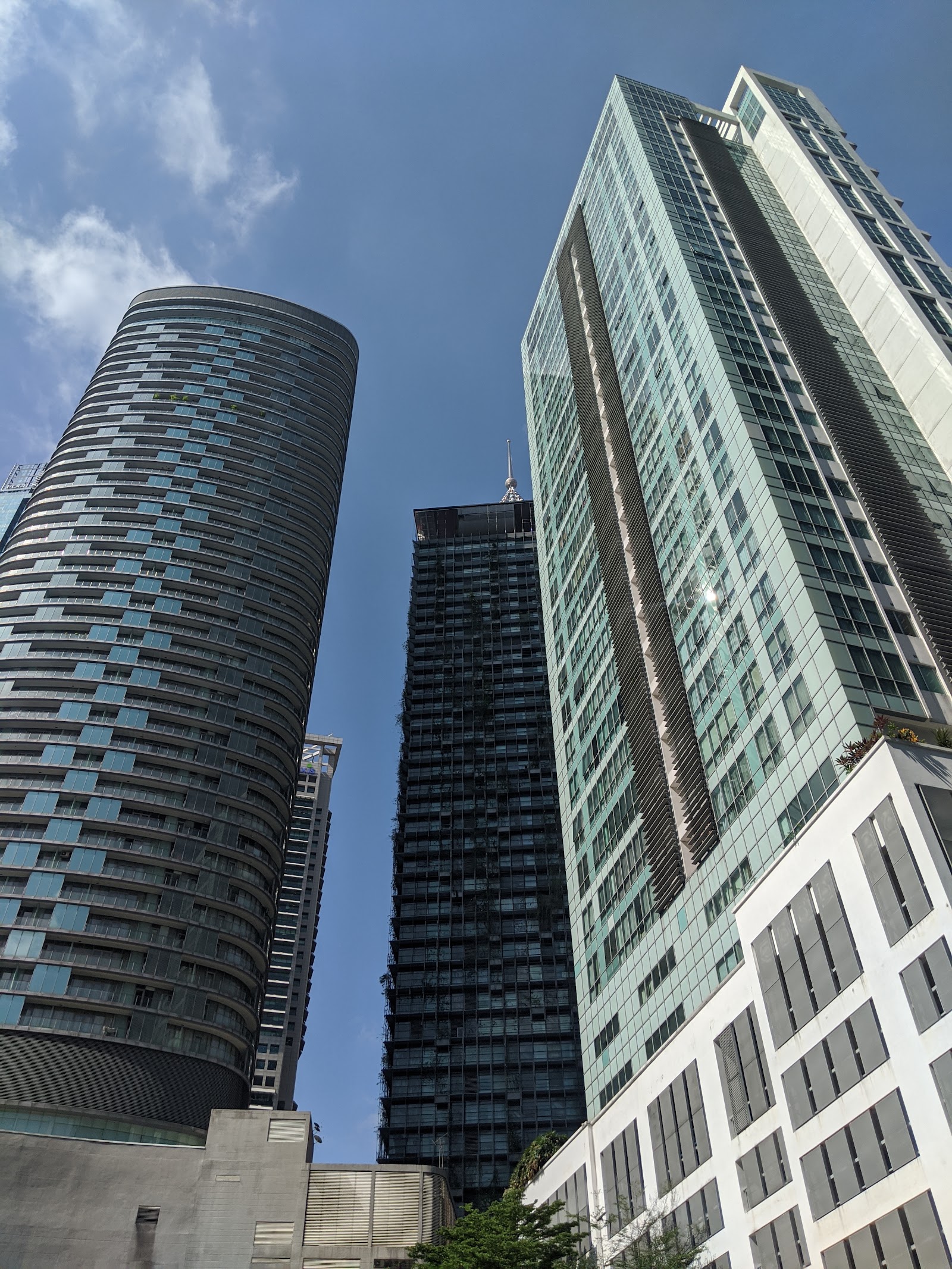 D'Mayang: A low-angle view of several high-rise buildings, including a cylindrical tower on the left, a dark green tower with visible vegetation in the center, and a modern glass tower on the right.