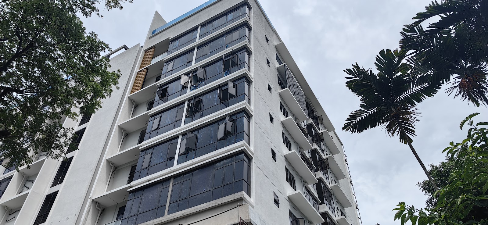 Impression U-Thant: A low-angle shot of a multi-story white apartment building with dark windows, framed by a tree on the left and a palm tree on the right against a cloudy sky.