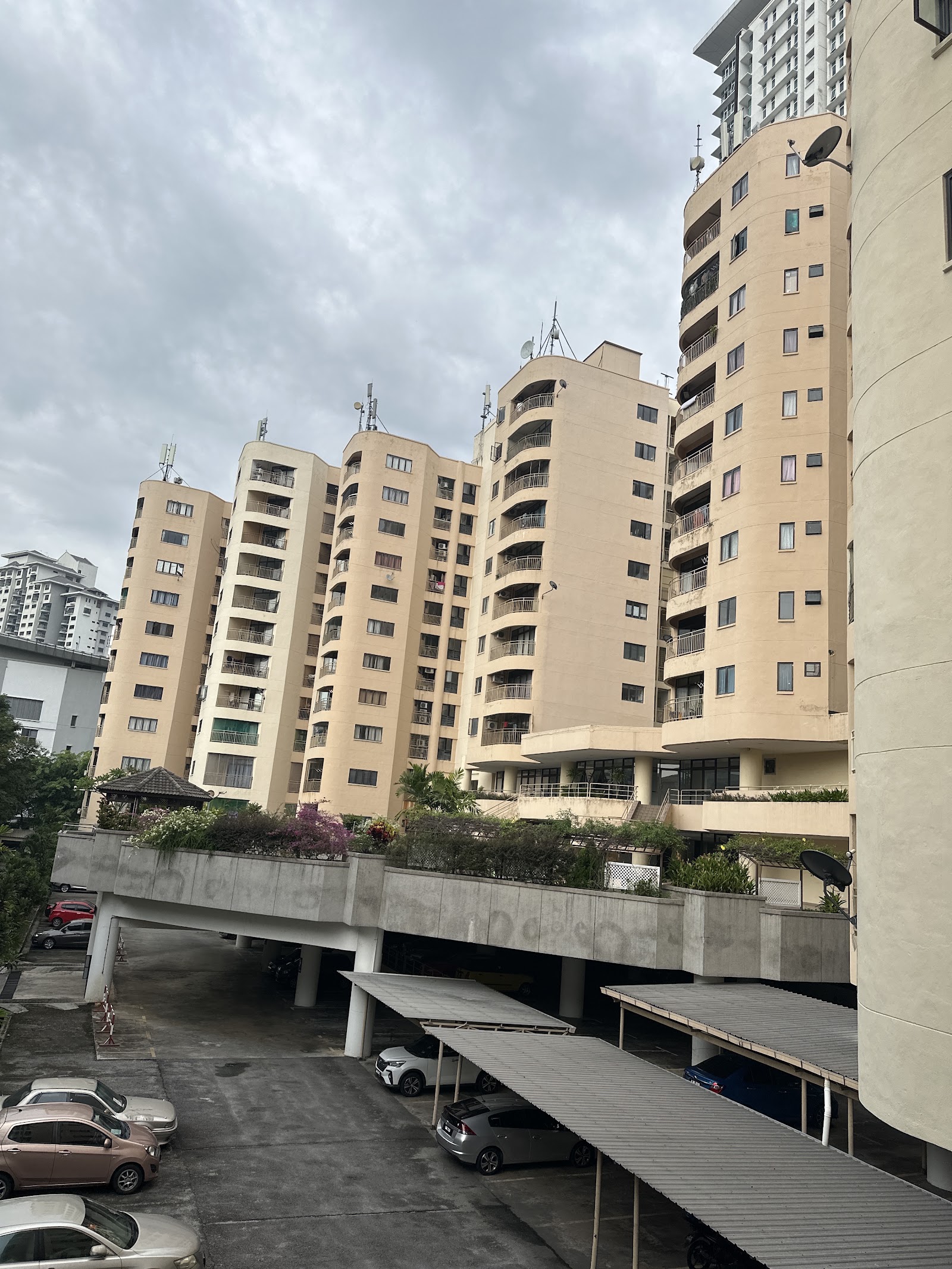 Indah Villa: A low-angle exterior view of several beige residential towers with curved balconies, standing over a parking area with covered and open stalls.