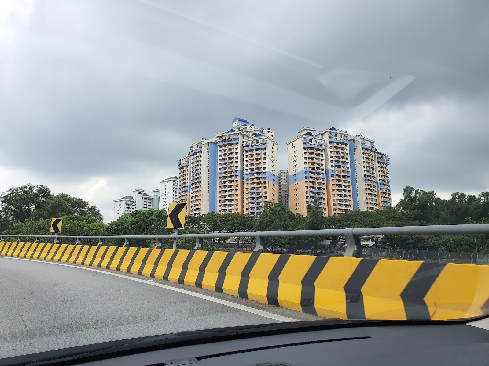 Vista Komanwel: A view of the Vista Komanwel residential towers from a road with a yellow and black chevron barrier, taken under a cloudy sky.