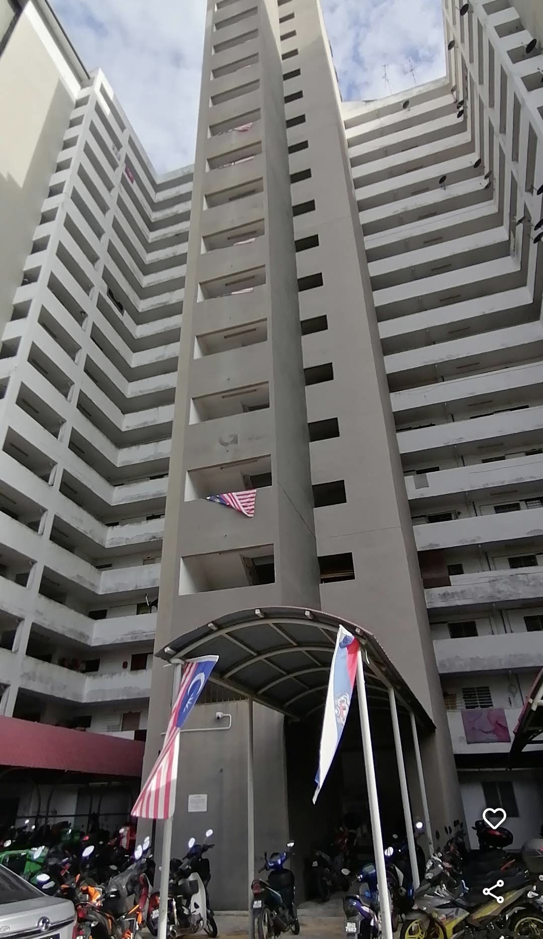 Apartment Seri Ceria 1: A low-angle view of the facade of a tall residential apartment building, showing the exterior of a central concrete tower and flanking wing sections, with a covered motorcycle parking area and entrance at the ground level.