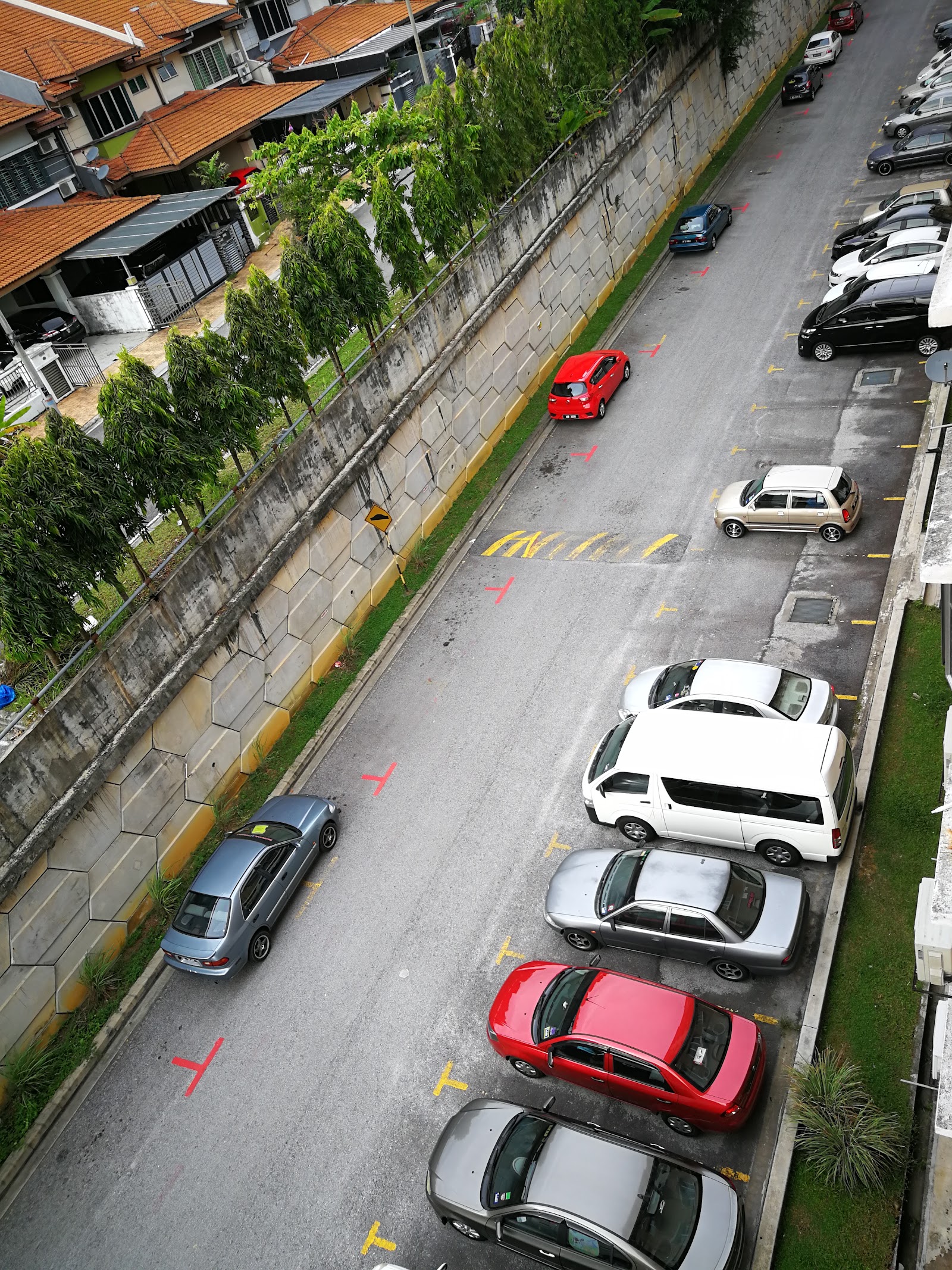 Pangsapuri Tainia: Aerial view of the residential development's parking area and external grounds, showing cars parked along a paved access road next to a retaining wall and a row of houses.