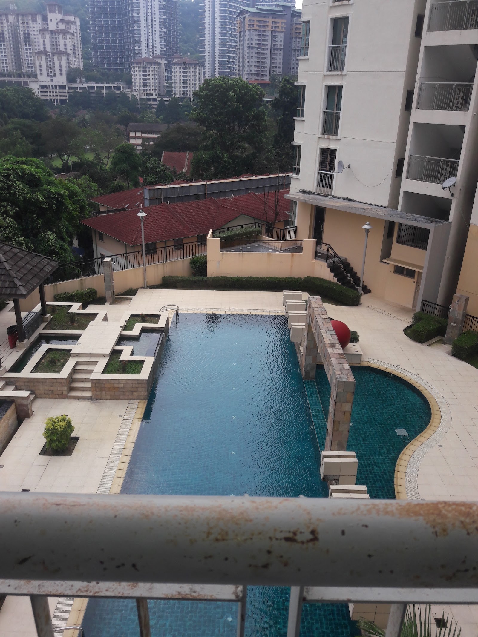 Ritze Perdana: A swimming pool area with a long main pool and a curved lap pool, surrounded by paved decks and landscaping, viewed from an elevated balcony.
