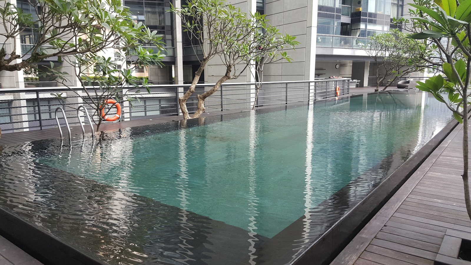 The Troika: An outdoor swimming pool with a ladder and safety rings surrounded by trees and a wooden deck, set in front of a modern apartment building.
