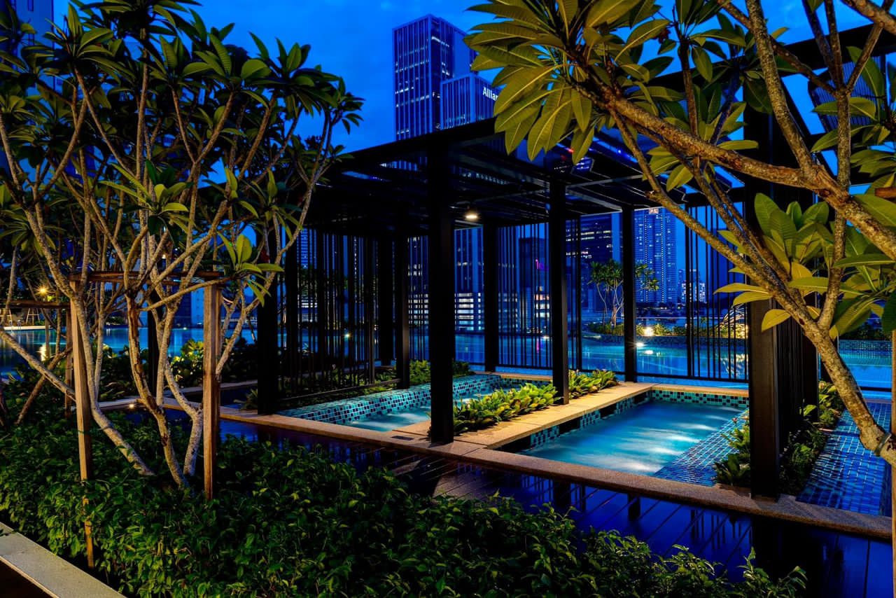 Sentral Suites: A serene nighttime view of a modern, landscaped rooftop pool area with illuminated water features, framed by trees and overlooking a glowing city skyline.
