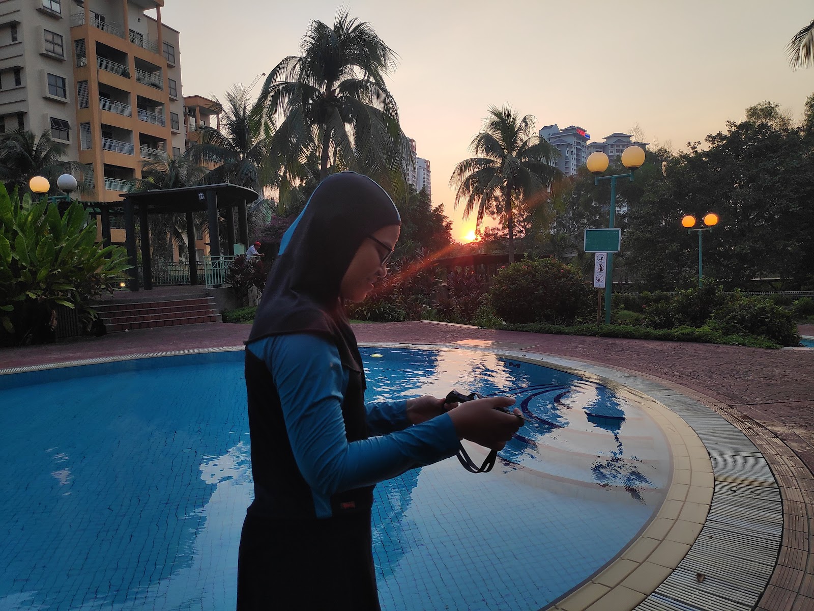 PALMVILLE RESORT CONDOMINIUM: A woman in a hijab looking at a phone stands beside a swimming pool at dusk, with resort buildings and palm trees in the background.
