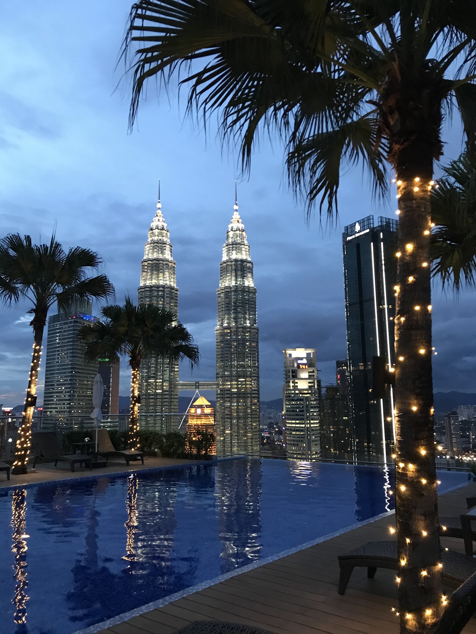 Quadro Residences: A rooftop infinity pool at night with palm trees and lounge chairs, overlooking the illuminated Petronas Twin Towers and surrounding city skyline.