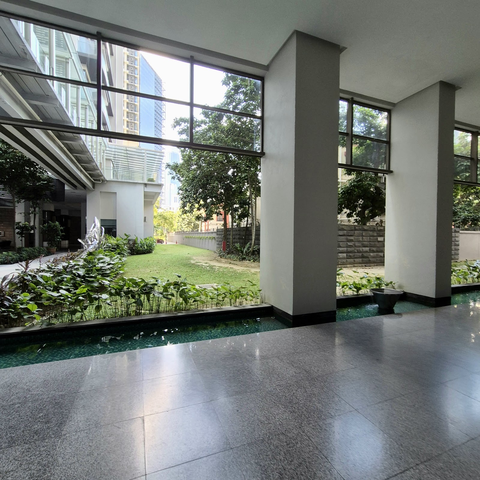 Suria Stonor: A covered common area with large pillars and floor-to-ceiling windows looking out onto a landscaped garden with a water feature and lawn.