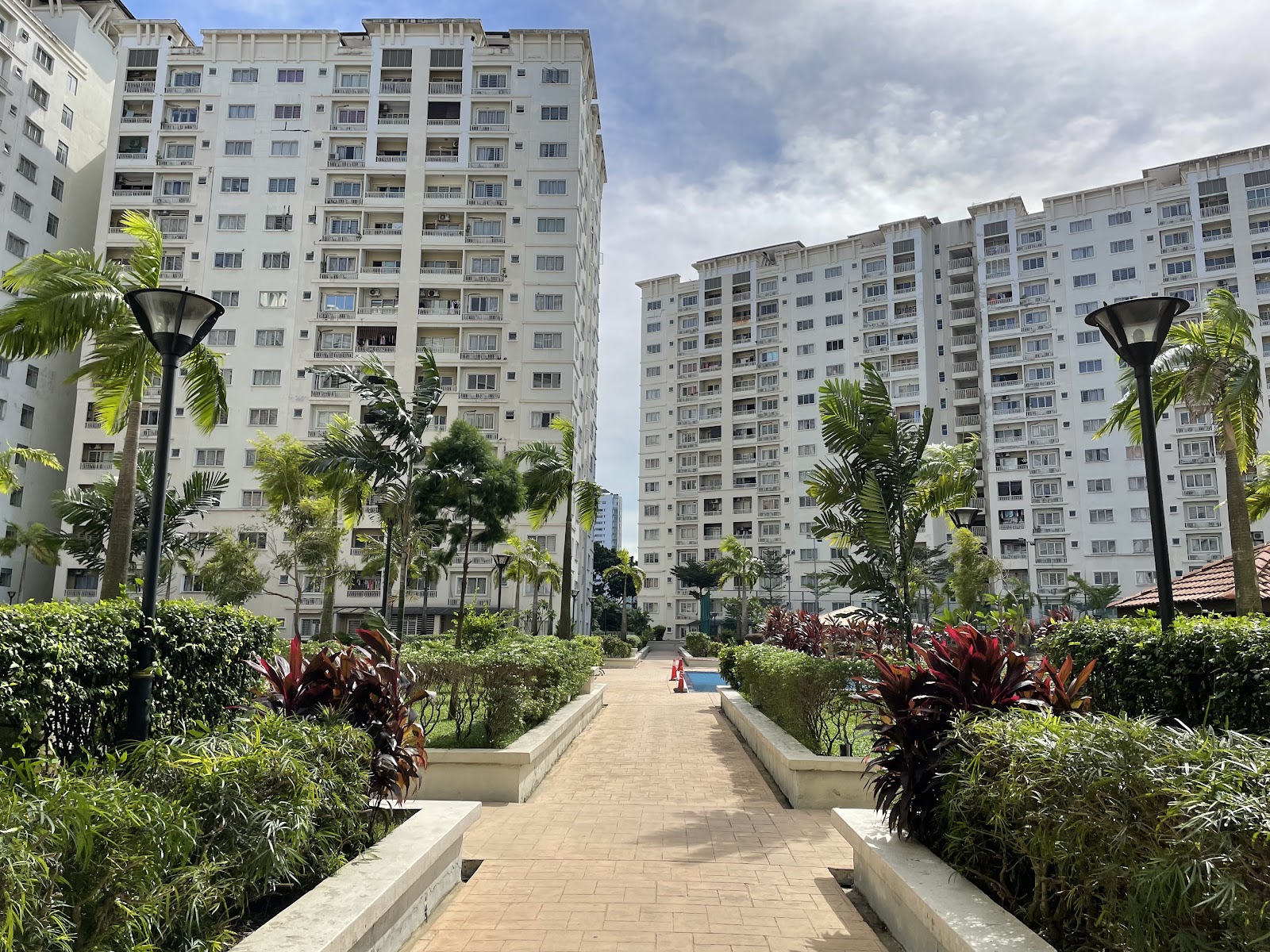 Suriamas: A landscaped pathway leading through the grounds of a residential development, flanked by tropical plants and lampposts with high-rise apartment towers in the background.