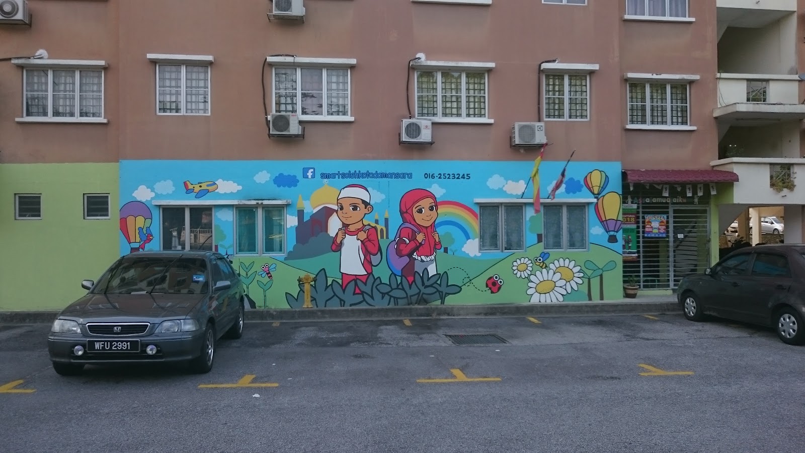 Pangsapuri Tainia: A wide shot of the lower exterior facade of a residential building featuring a colorful mural of children and a parking lot with cars.