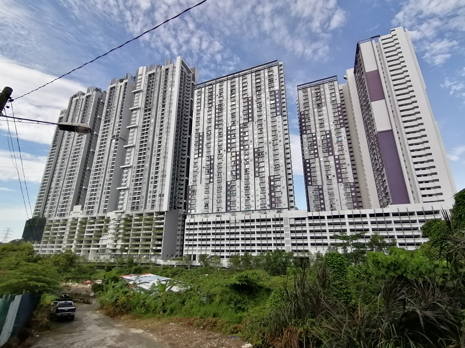Residensi Jalilmas @ Bukit Jalil: A low-angle exterior view of high-rise residential towers rising above a foreground of overgrown green vegetation and a dirt path.