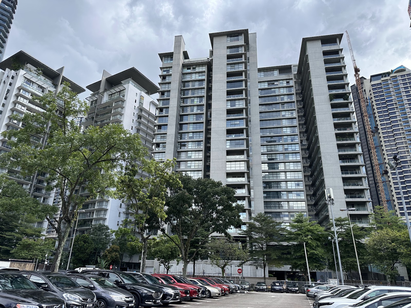 Park Seven: A row of parked cars in the foreground, with a line of trees and several high-rise apartment buildings in the background under a cloudy sky.