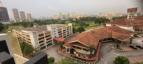 The Tropika Bukit Jalil: Elevated view of the Tropika Bukit Jalil complex, showing a multi-story car park and main building with a tiled roof, set against a backdrop of green fields and high-rise apartments.