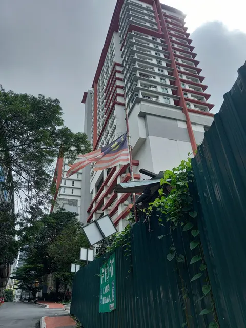 Ritze Perdana 2: A view of the facade of a tall residential building with white walls and red trim, featuring balconies and air conditioning units, with a Malaysian flag flying on a pole and greenery in the foreground.