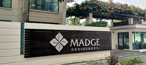 Madge Residences: An entrance sign for Madge Residences mounted on a white wall, with a paved driveway, a security guard house, and flowering vines on a pergola in the background.