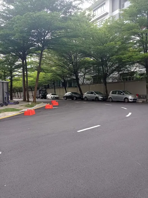 Madge Mansions Condominiums: A paved driveway and car park with several parked vehicles, including a silver hatchback and a black sedan, set among trees with a multi-story building visible in the background.