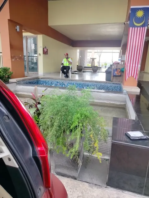 Ritze Perdana: A security guard in a neon vest sits on a chair near a small tiled swimming pool and plants at the entrance of a building, with a Malaysian flag hanging nearby.
