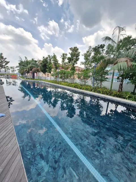 YOLO Signature Suites: A long, modern swimming pool with a wooden deck, surrounded by lush greenery and palm trees, reflecting the cloudy sky.